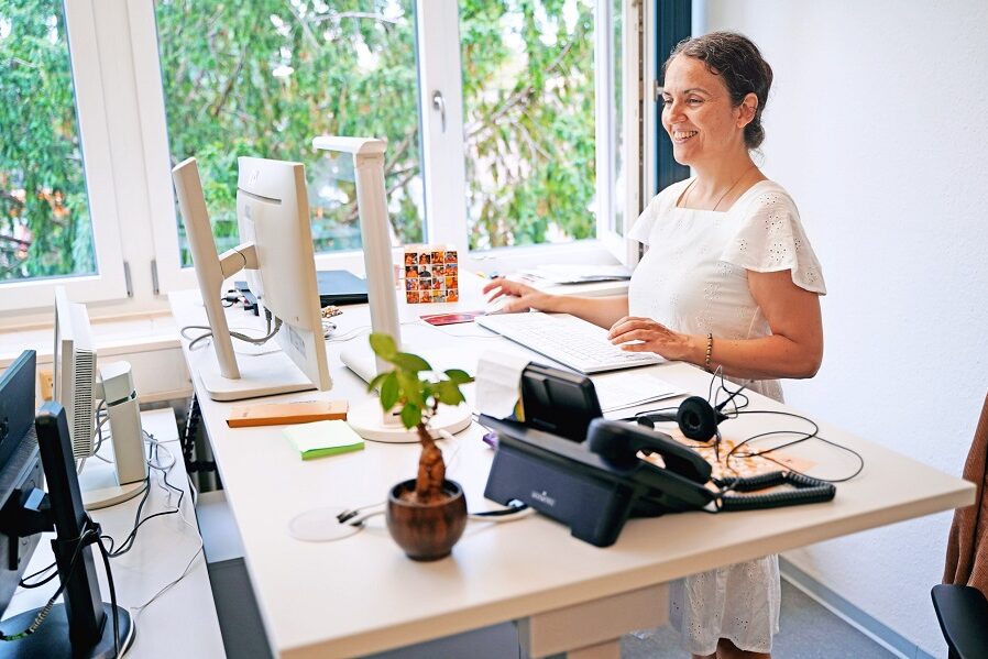 Eine Person steht lächelnd an einem Schreibtisch im Büro und schaut auf einen Bildschirm.