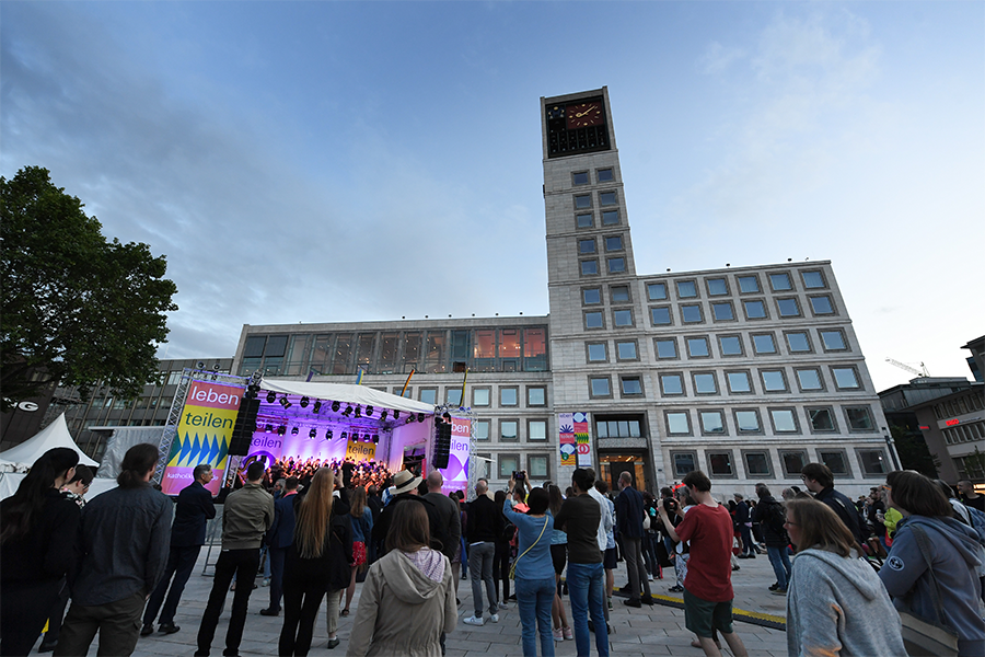Menschen stehen vor einer Bühne mit Scheinwerfern, im Hintergrund das Rathaus
