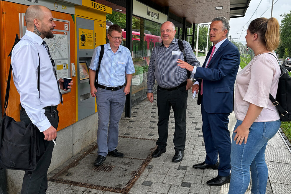 Vier Männer und eine Frau stehen an einer Haltestelle und unterhalten sich.