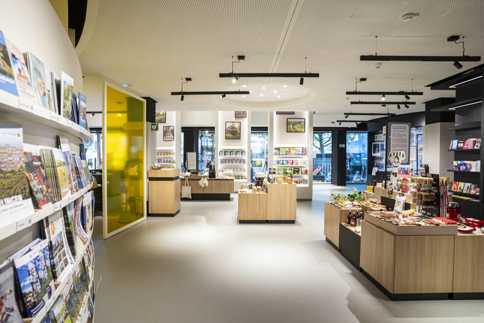 Die moderne Tourist‐Information i‐Punkt im Erdgeschoss des Hauses des Tourismus in Stuttgart. In Wandregalen sind Informationsflyer ausgestellt. Auf Regalen in der Raummitte werden Souvenirs und regionale Produkte präsentiert.