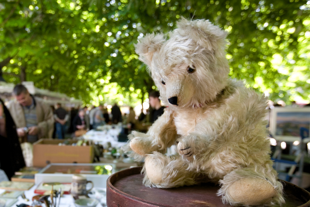 Teddybär sitzt auf einer kleinen Tisch, im Hintergrund ist ein Flohmarkt zu sehen.