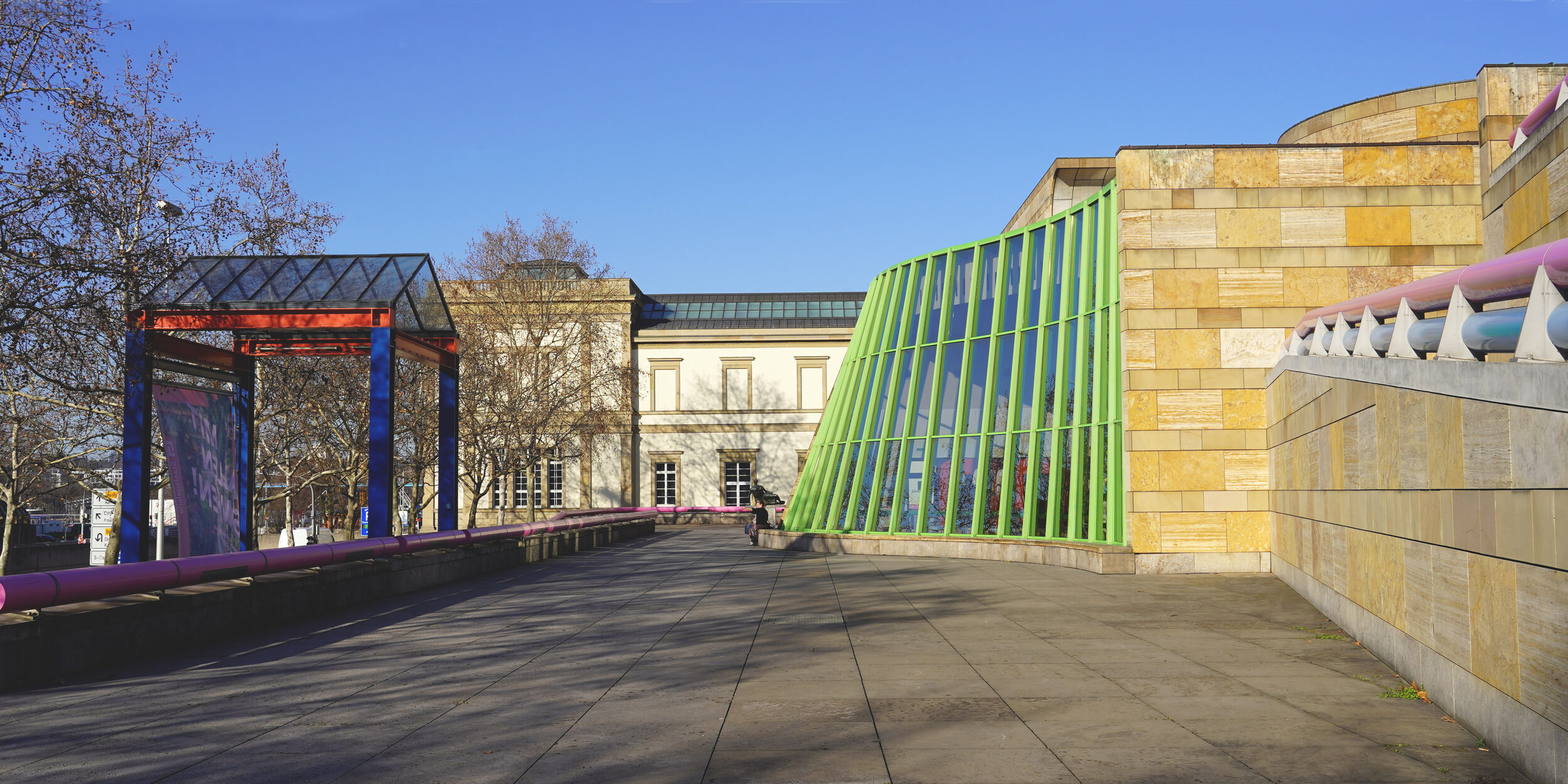 Neue Staatsgalerie mit geschwungener Travertin-Fassade, mit pinken und blauen Handläufen an den Treppen sowie grünen Stahlträgern.
