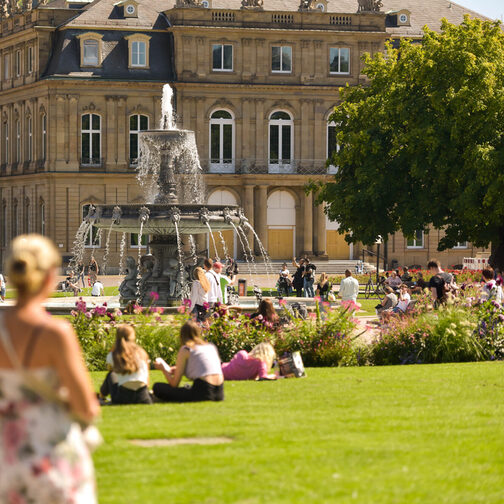 Am Schlossplatz sitzen Menschen bei Sonnenschein auf der Wiese und am Brunnen.