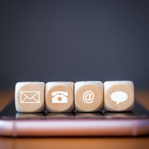 Drei hölzerne Würfelblöcke auf einem Smartphone. Auf jedem Würfel ist ein Symbol abgebildet: Telefon, Adresse, E-Mail, Sprechblase