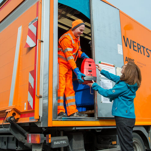 Ein Mann steht in einem orangenen Lastwagen und nimmt einer Frau einen Staubsauger ab.