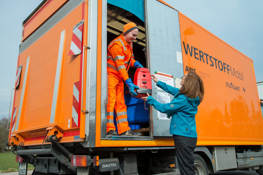 Ein Mann steht in einem orangenen Lastwagen und nimmt einer Frau einen Staubsauger ab.