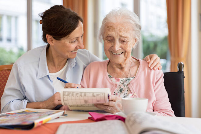 Eine Seniorin löst ein Kreuzworträtsel, eine Frau sitzt neben ihr und legt ihr den Arm um die Schulter.