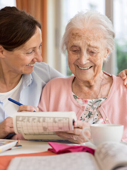 Eine Seniorin löst ein Kreuzworträtsel, eine Frau sitzt neben ihr und legt ihr den Arm um die Schulter.