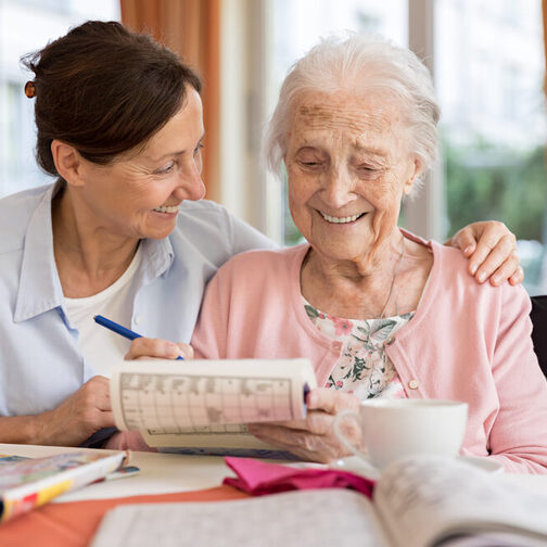 Eine Seniorin löst ein Kreuzworträtsel, eine Frau sitzt neben ihr und legt ihr den Arm um die Schulter.