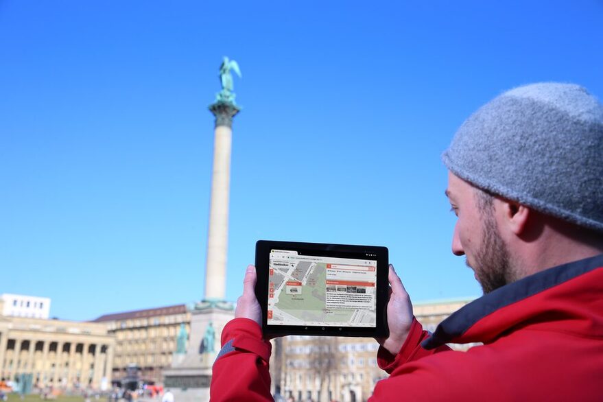 Ein Mann, rechts im Bild, hält ein Tablet hoch, das ihm Informationen zum Stuttgarter Schlossplatz anzeigt, der im Hintergrund zu sehen ist.