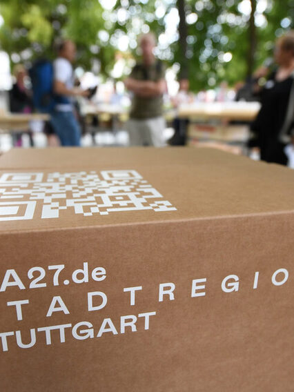 Ein Karton mit der Aufschrift "IBA27.de, Stadtregion Stuttgart". Im Hintergrund Menschen im Gespräch.