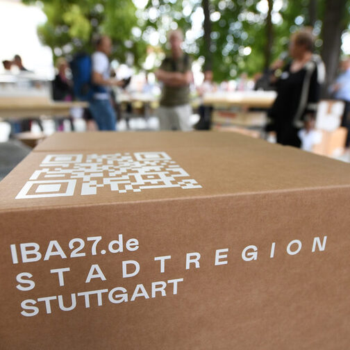 Ein Karton mit der Aufschrift "IBA27.de, Stadtregion Stuttgart". Im Hintergrund Menschen im Gespräch.