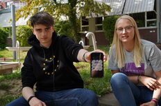 Zwei junge Menschen sitzen in einem Park, eine Person haält ein Smartphone in die Kamera.