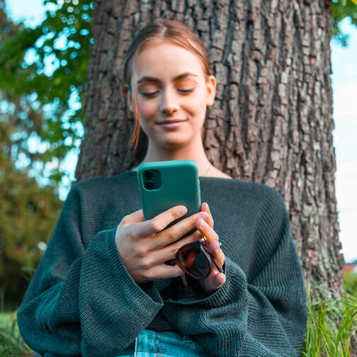 Jugendliche lehnt am Baum, schaut auf ihr Smartphone und lächelt zufrieden