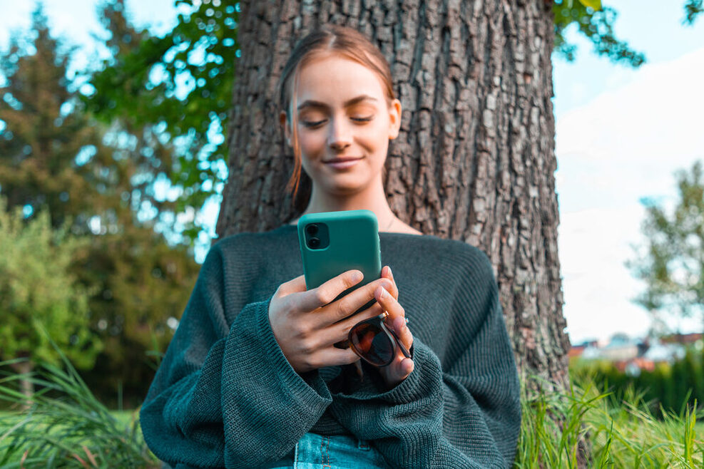Jugendliche lehnt am Baum, schaut auf ihr Smartphone und lächelt zufrieden