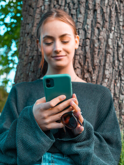 Jugendliche lehnt am Baum, schaut auf ihr Smartphone und lächelt zufrieden