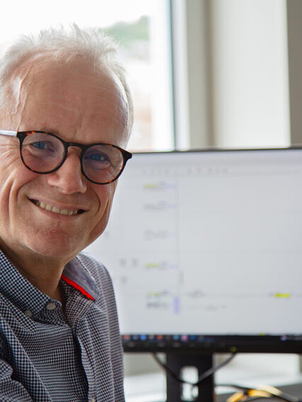 Ein männlicher Mitarbeiter dreht sich lächelnd am Schreibtisch vor Computerbildschirmen zur Kamera hin.