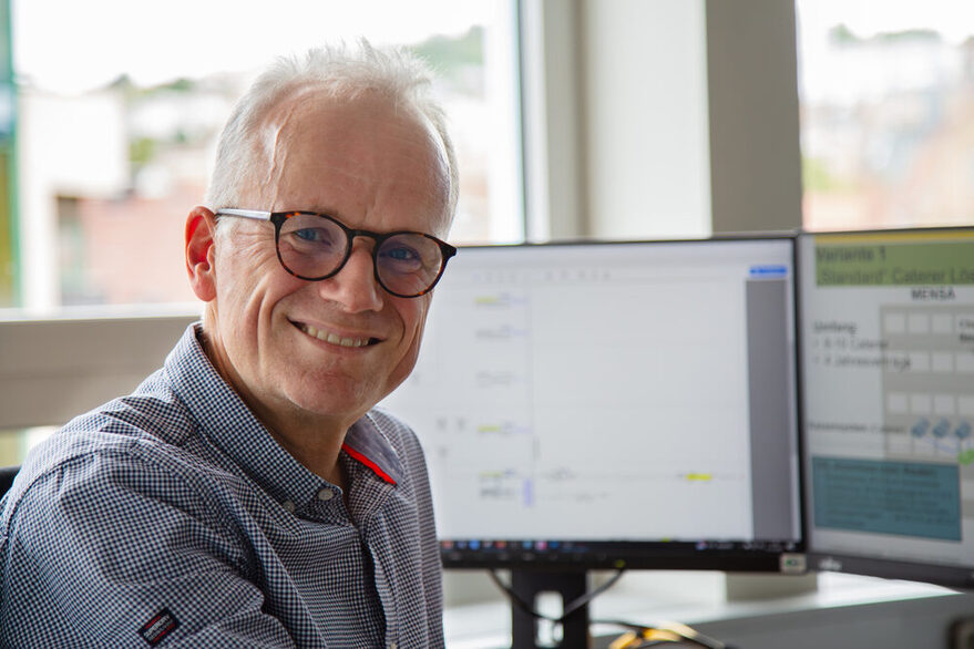 Ein männlicher Mitarbeiter dreht sich lächelnd am Schreibtisch vor Computerbildschirmen zur Kamera hin.