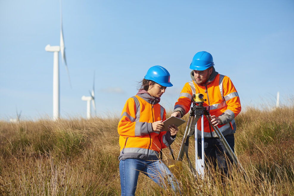 Zwei Arbeiter in orangen Jacken stehen in einem Windpark mit einem Vermessungsgerät.