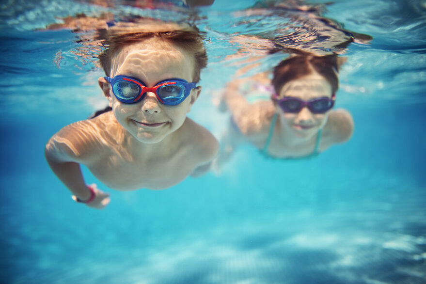 Ein Junge im Vordergrund und ein Mädchen im Hintergrund tauchen in einem Schwimmbecken auf die Kamera zu.