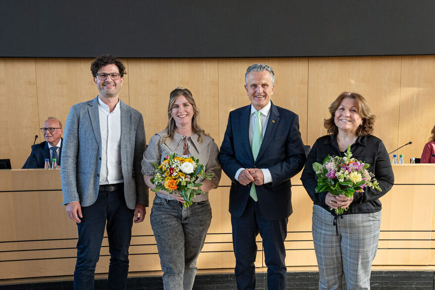 Zwei Frauen und zwei Männer stehen nebeneinander und blicken in die Kamera. Die Frauen halten Blumensträuße in den Händen.