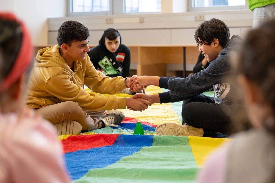 Zwei Jugendliche sitzen in einem Sitzkreis und geben sich die Hände.