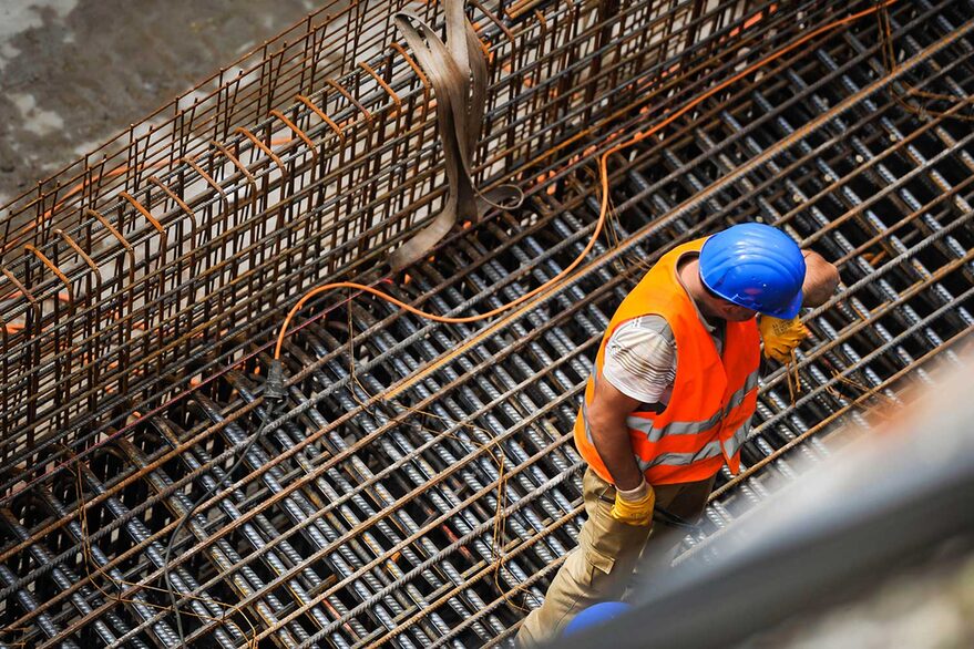 Zu sehen ist ein Bauarbeiter, der auf einem Gerüst einer größeren Baustelle steht. Er hat einen blauen Helm und eine orangefarbene Warnweste an. Das Bild wurde von schräg oben aufgenommen.