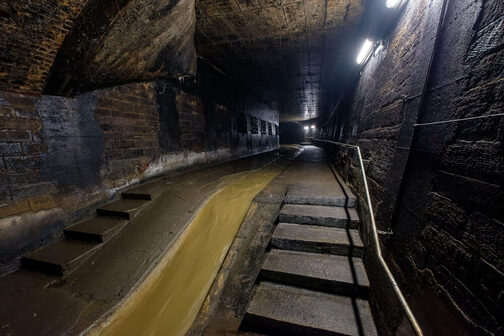 Auf dem Foto ist der Abwasserkanal zu sehen, an den Seiten gibt es einen Gehtsteig mit Treppen und einem Geländer an der rechten Wand.
