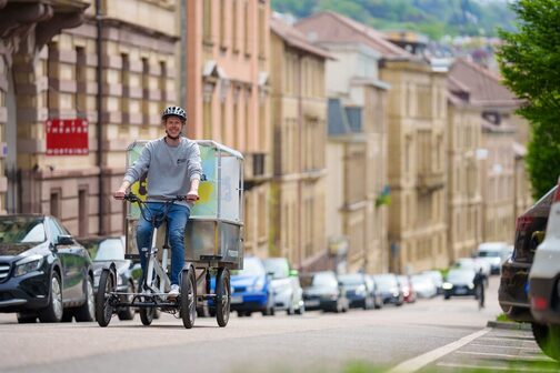 A young man rides a cargo bike through a Stuttgart street in a good mood. The buildings in the background are blurred, the man and the bike are in focus.