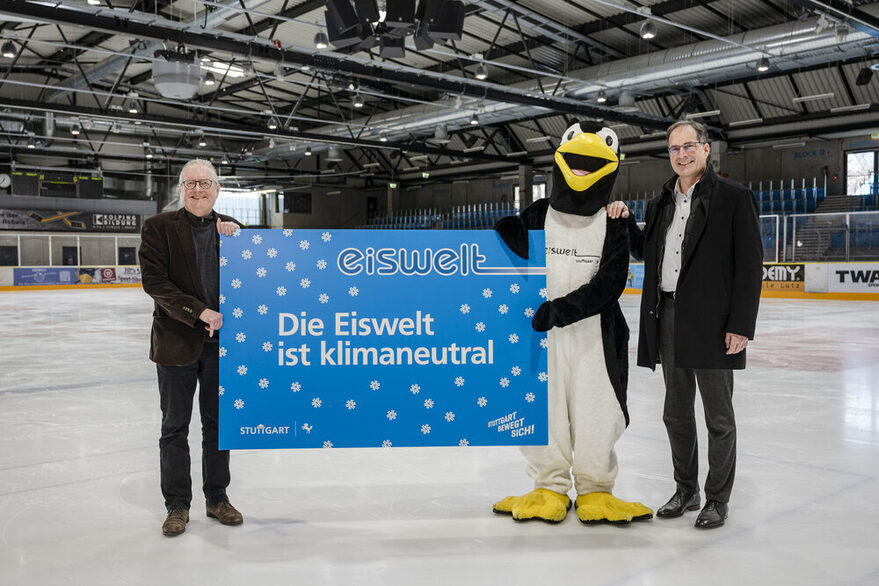 Die Bürgermeister Peter Pätzold und Dr. Clemens Maier mit dem Masskotchen der Eiswelt, Pinguin Fridolin Frostig, auf dem Eis in der Eiswelt mit einem Schild: "Die Eiswelt ist klimaneutral."