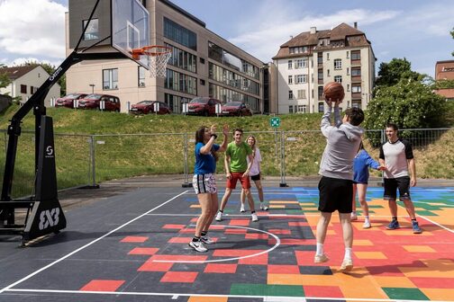 Sechs Jugendliche spielen begeistert Basketball auf einem farbenfrohen Outdoor-Basketballfeld, umgeben von urbaner Kulisse.