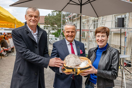 Drei Personen halten einen Korb mit Brot und Salz, im Hintergrund eine Baustelle.