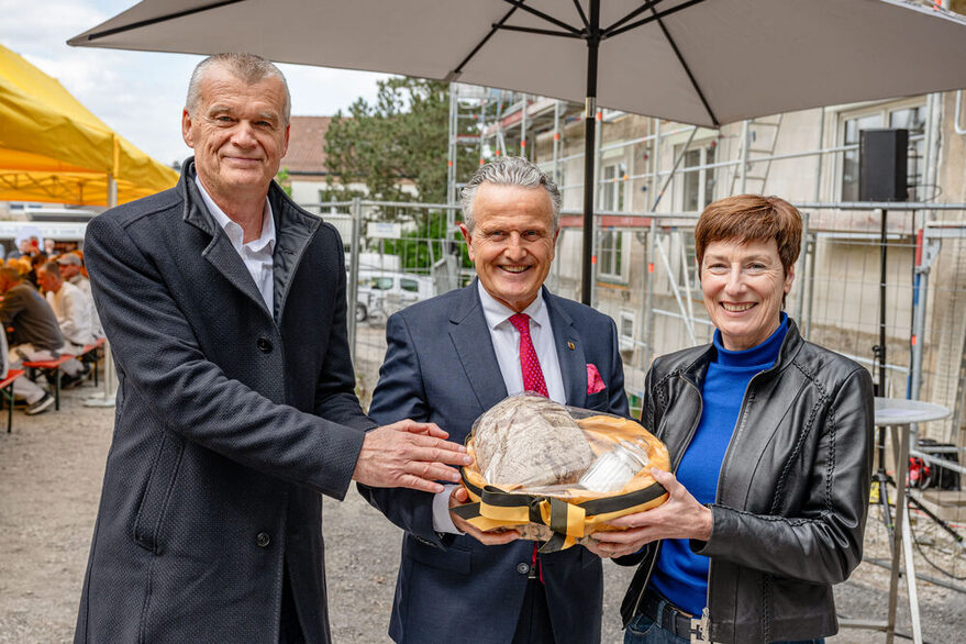 Drei Personen halten einen Korb mit Brot und Salz, im Hintergrund eine Baustelle.