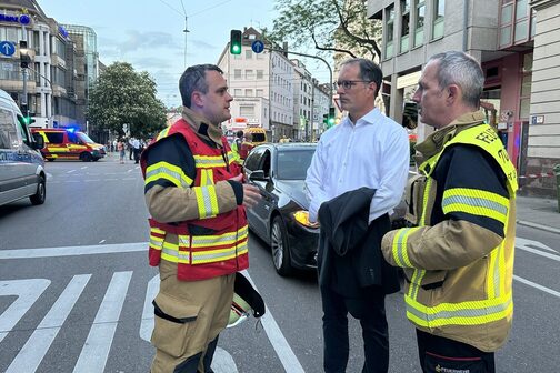 Zwei Feuerwehrmänner und ein Mann im Anzug stehen auf der Straße und unterhalten sich.