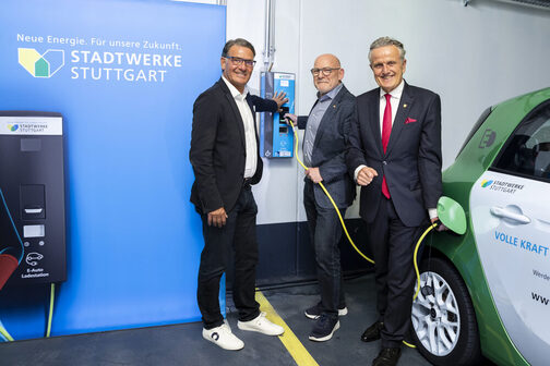 Three men are standing in front of an electric charging station in a parking garage, charging a car.