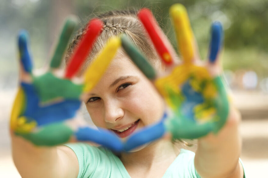 Ein lächelndes Mädchen hält seine mit bunter Farbe bemalten Handflächen in die Kamera.