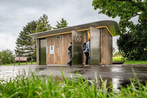 Eine moderne und barrierefreie Toilettenanlage in der Natur in Stuttgart-Rotenberg. Außenrum sind Bäume, zwei Menschen gehen in die Toilette rein.