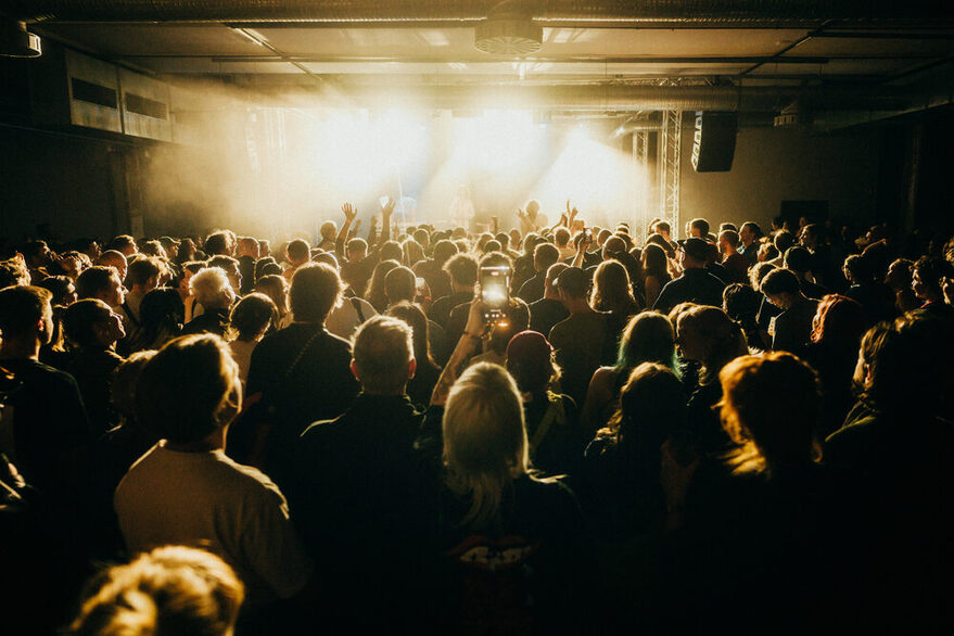 Viele Menschen stehen als Publikum versammelt vor einer lichtdurchfluteten Bühne und feiern.