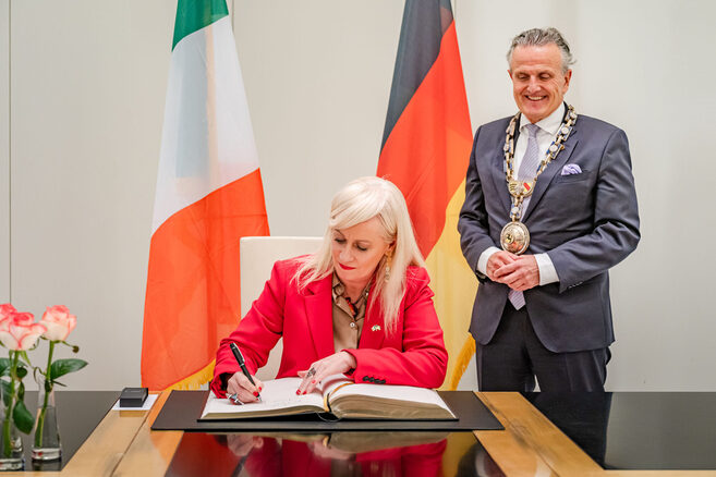 Eine Frau schreibt in ein großes Buch. Ein Mann steht daneben und schaut ihr zu.