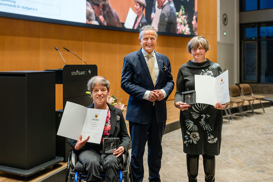 Ein Mann und zwei Frauen schauen in die Kamera. Die Frauen halten Urkunden und Ehrenplaketten in den Händen. Eine der Frauen sitzt im Rollstuhl.