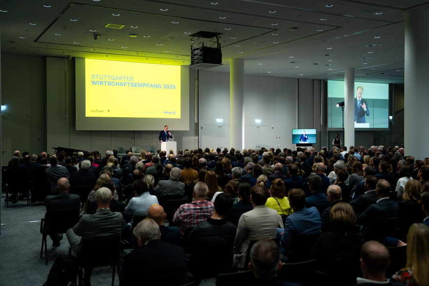 Die Gäste des 3. Stuttgarter Wirtschaftsempfangs sitzen in einem abgedunkelten Raum im Publikum und hören der Eröffnungsrede auf dem Podium zu.