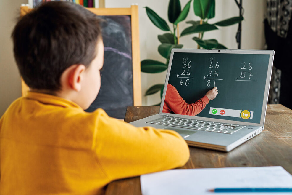 Ein Grundschüler sitzt vor einem Lapotop. Auf dem Bildschirm macht ein Lehrer Rechenaufgaben.
