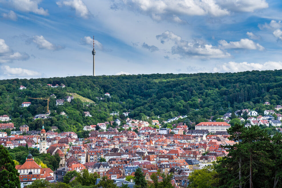 Stadtpanorama von Stuttgart: Häuser mit roten Dächern in einem Tal, umgeben von grünem Wald, auf einem Hügel steht der Fernsehturm.