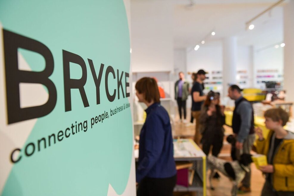 Logo von Brycke, im Hintergrund ist der Pop-up-Space mit vielen Besuchern sichtbar.