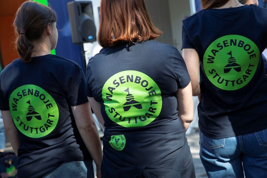 Drei junge Frauen stehen in einer Reihe und sind von hinten fotografiert. Alle drei tragen ein T-Shirt mit dem großen Aufdruck "Wasenboje Stuttgart".