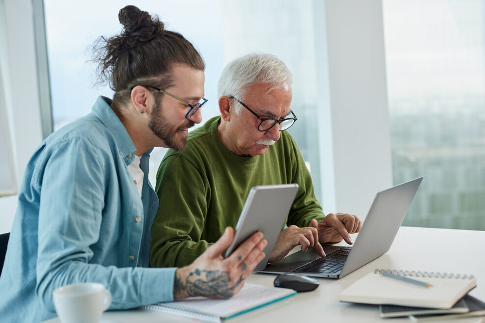 Ein jüngerer Mann und ein älterer Mann vor einem Tablet und einem Computer.