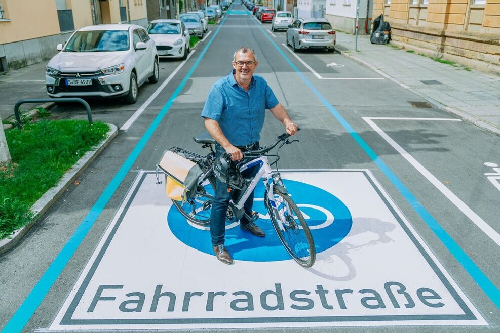 Jürgen Mutz stellte die Fahrradstraße vor Ort vor. Mutz hat ein Fahrrad stehend bemannt und lächelt in die KAmera. Er steht auf einem neuen Piktogramm mit dem Hinweis auf die Fahrradstraße.