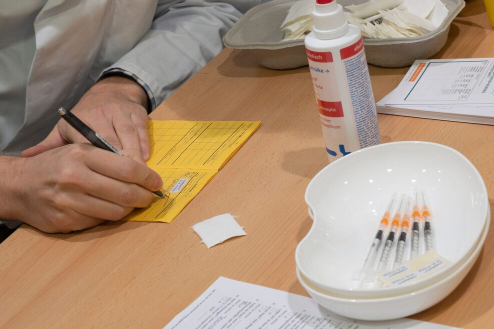 Eine Person trägt in einen gelben Impfpass das Datum einer Impfung ein. Auf dem Tisch liegen in einer Schale aufgezogene Spritzen, daneben steht ein Desinfektionsmittel.