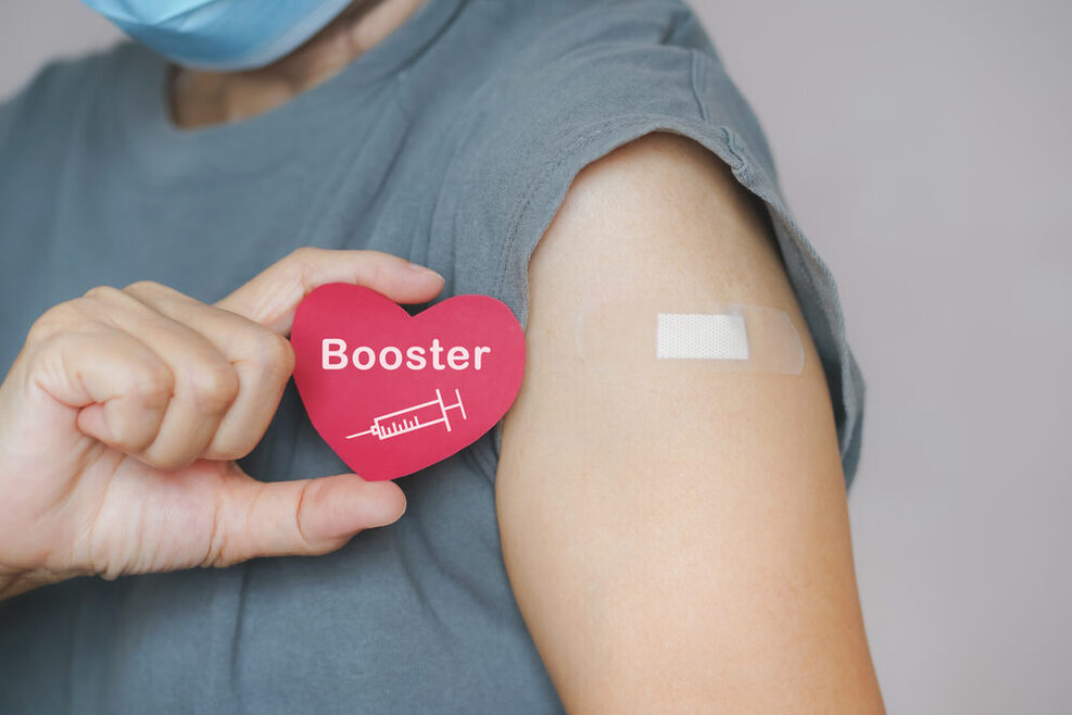 Eine Person zeigt eine rote Herzform mit Spritzensymbol und den Worten Booster. Auf dem Oberarm klebt ein Pflaster nach einer Impfung.