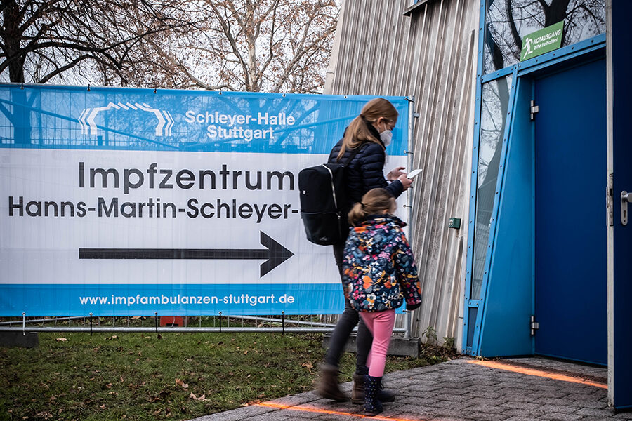 Eine Mutter geht mit ihrem Kind auf einen Eingang rechts zu. Im Hintergrund ist ein Banner mit der Aufschrift "Impfzentrum Hanns-Martin-Schleyer-Halle" zu sehen.